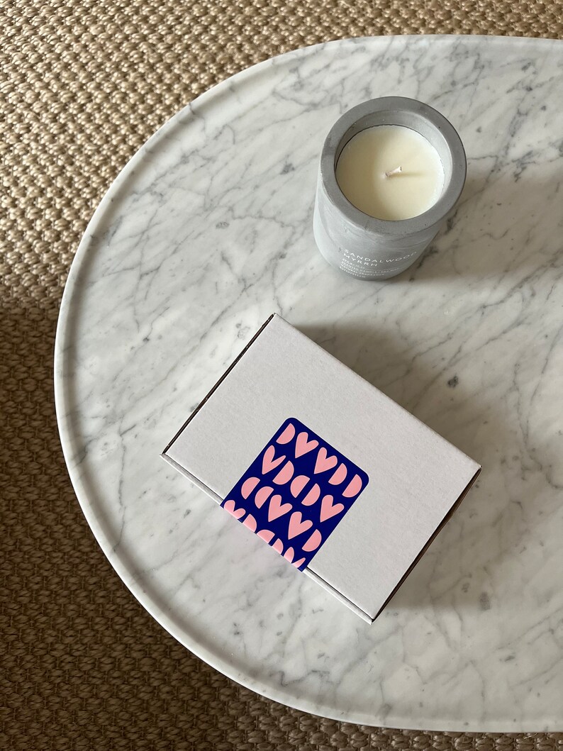 May include: A white candle in a gray concrete jar with the text "Sandalwood Myrrh" on the side. The candle is sitting on a white marble surface with a white box with a pink and blue heart pattern on top.