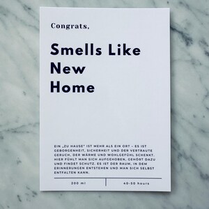 May include: White card with black text that reads "Congrats, Smells Like New Home". The card also includes the text "Ein zu Hause' ist mehr als ein Ort - es ist Geborgenheint, Sicherheit und der vertraute Geruch, der W&auml;rme und Wohlgef&uuml;hl schenkt. Hier f&uuml;hlt man sich aufgehoben, geh&ouml;rt dazu und findet Schutz. Es ist der Raum, in dem Erinnerungen entstehen und man sich selbst entfalten kann."  The card also includes the text "200 ml" and "40-50 hours".