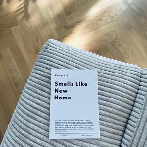 May include: A white card with the text "Congrats. Smells Like New Home" on a light gray corduroy surface. The card also includes the text "Ein zu Hause ist mehr als ein Ort - es ist Geborgenheint, Sicherheit und der vertraute Geruch, der warme und wohlgefuhl schenkt. Hier fuhlt man sich aufgehoben, gehoert dazu und findet Schutz. Es ist der Raum, in dem Erinnerungen entstehen und man sich selbst entfalten kann. 200 ml 40-50 hours"