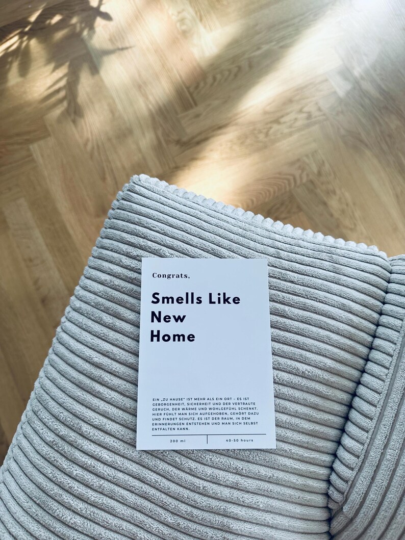 May include: A white card with the text "Congrats. Smells Like New Home" on a light gray corduroy surface. The card also includes the text "Ein zu Hause ist mehr als ein Ort - es ist Geborgenheint, Sicherheit und der vertraute Geruch, der warme und wohlgefuhl schenkt. Hier fuhlt man sich aufgehoben, gehoert dazu und findet Schutz. Es ist der Raum, in dem Erinnerungen entstehen und man sich selbst entfalten kann. 200 ml 40-50 hours"