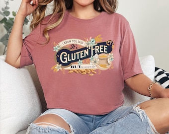 May include: A pink t-shirt with a vintage-style graphic that says "It's Gluten Free But... Can I just see the label?" The graphic features a wheat stalk, a flower, and a bag of flour.