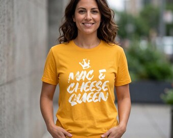 May include: A mustard yellow short-sleeved t-shirt featuring a white graphic print of a crown above the text "MAC & CHEESE QUEEN" in a bold, distressed font style. The shirt has a crew neckline and is made of a solid-colored cotton fabric.
