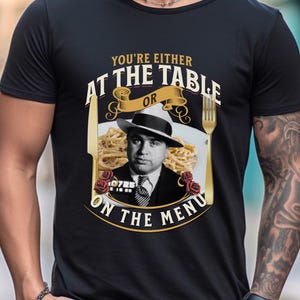 May include: A black t-shirt with a graphic design featuring a man in a fedora and a plate of spaghetti. The text on the shirt reads "You're either at the table or on the menu".