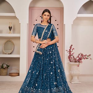 May include: A teal blue net lehenga with silver embroidery and sequins. The lehenga has a matching dupatta with a silver border. The model is wearing a matching teal blue blouse with a net neckline.