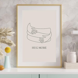 Op de afbeelding: Een ingelijste kunstdruk met een gouden lijst. Het kunstwerk toont een minimalistische lijntekening van twee armen die elkaar omarmen. De woorden "HUG MORE" zijn in zwart onder de tekening gedrukt. De achtergrond is lichtbeige.