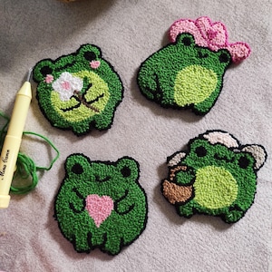 May include: Four green felt frog ornaments with different details. One frog is wearing a pink hat, one is holding a pink flower, one is holding a pink heart, and one is holding a brown basket.