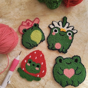 May include: Four green frog-shaped needle felted ornaments. One frog is wearing a pink flower crown, another has a white daisy on its head, one is inside a red strawberry, and the last one is holding a pink heart.