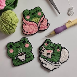 May include: Handmade frog-shaped embroidered patches in green, pink, and white. One frog is wrapped in a pink blanket, another holds a coffee cup, and the third reads a book. Green and pink yarn balls, scissors, and a punch needle are also visible.