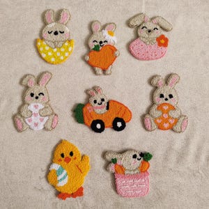 May include: Nine felt bunny and chick ornaments in various poses. The bunnies are holding carrots, hearts, and eggs. The chick is holding a green and white striped egg. The ornaments are all in pastel colors.
