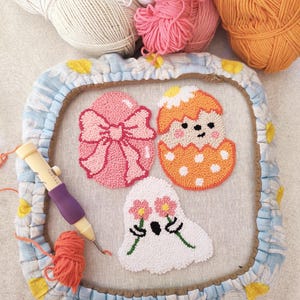 May include: A punch needle embroidery kit with three completed designs: a pink egg with a bow, an orange egg with a face, and a white ghost with flowers. The kit includes a punch needle, yarn, and a hoop.