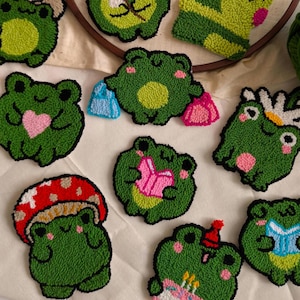 May include: A collection of ten colorful, hand-stitched felt frog ornaments. The frogs are various shades of green and feature different details, such as hearts, flowers, and books.
