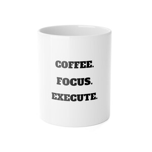 Könnte beinhalten: Weißer Keramikbecher mit den Worten "COFFEE. FOCUS. EXECUTE." in Schwarz vertikal aufgedruckt. Der Becher hat eine einfache, zylindrische Form und einen Henkel auf der rechten Seite.