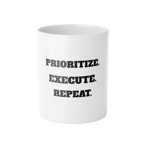 May include: A white ceramic mug with the words "PRIORITIZE. EXECUTE. REPEAT." printed in bold, black capital letters. The mug is cylindrical and appears to be empty.