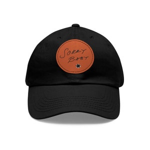 May include: Black baseball cap with a brown circular patch. The patch features the handwritten text "Sorry Baby" in black script. A small, white star-shaped design is below the text. The cap is made of a soft material.