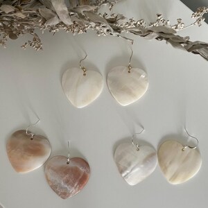 Peut inclure: Trois paires de boucles d'oreilles en forme de cœur fabriquées en nacre irisée. Chaque paire présente une variation de couleur distincte, incluant le blanc crème, le pêche doux et un mélange de tons brun clair et blanc, suspendues à des crochets.