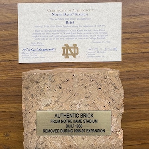 May include: An authentic brick from Notre Dame Stadium, built in 1930 and removed during the 1996-97 expansion. Includes a certificate of authenticity. The brick is a light brown color and has a metal plaque.