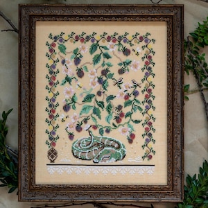 May include: A cross-stitch embroidery framed in a dark brown wood frame. The design features a green snake curled up under a flowering bush with white blossoms and black berries.