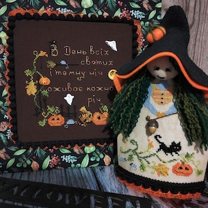 Op de afbeelding: Een handgemaakte Halloween-heksenpop met een zwarte hoed, oranje rand en groen garenhaar. De pop draagt een witte jurk met oranje en blauwe accenten en houdt een kleine eikel vast. De pop zit voor een bruine stof met een kruissteekontwerp waarop "В День всіх святих і тому чи оживає кожно річ" staat.