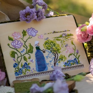 Op de afbeelding: Kruissteekborduurwerk met een vrouw in een blauwe jurk die een tuin met bloemen water geeft. De borduursel is omlijst met een groene rand en omgeven door paarse en roze bloemen. De scène toont een serene tuinlandschap.