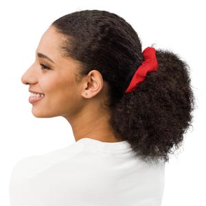 Puede incluir: Una mujer con cabello oscuro recogido en una cola de caballo, asegurada con una colorida y roja goma para el pelo. Lleva una camiseta blanca y sonríe. El fondo es blanco.