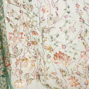 a close-up view of a fabric with a floral pattern, showcasing a variety of colorful flowers and leaves. The fabric appears to be made of a light-colored material, possibly cotton or linen, and is adorned with numerous small, colorful beads or sequins that add texture and visual interest to the design. The intricate details and vibrant colors of the floral pattern make the fabric a visually appealing and eye-catching piece.