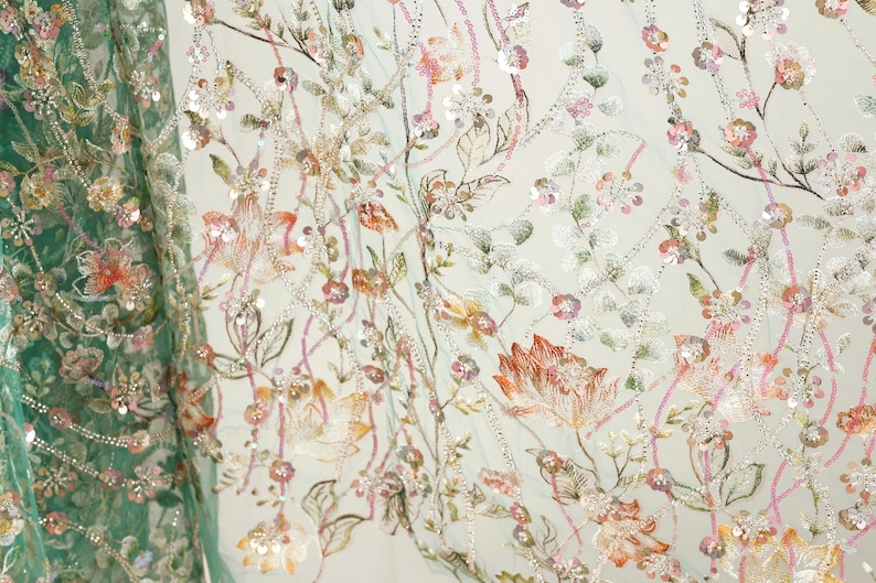 a close-up view of a fabric with a floral pattern, showcasing a variety of colorful flowers and leaves. The fabric appears to be made of a light-colored material, possibly cotton or linen, and is adorned with numerous small, colorful beads or sequins that add texture and visual interest to the design. The intricate details and vibrant colors of the floral pattern make the fabric a visually appealing and eye-catching piece.