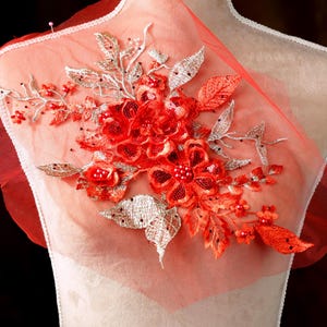Puede incluir: Aplicación floral con flores y hojas bordadas en rojo y plata sobre una tela transparente color melocotón. La aplicación está adherida a un maniquí, mostrando su uso potencial en proyectos de moda o artesanía.