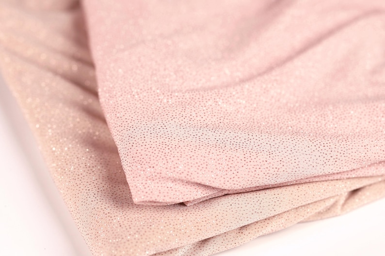 A close-up image of a fabric with a pink or peach-colored texture, featuring small, glittery dots or specks that create a sparkling effect.