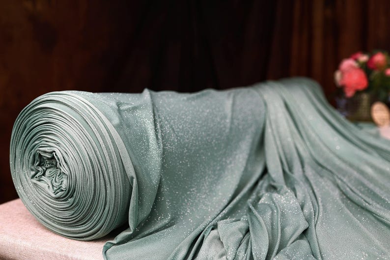 A close-up view of a roll of shimmering blue fabric with a floral pattern, placed on a table against a dark background.