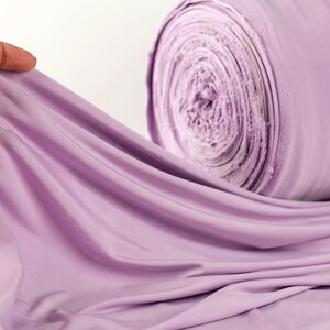 A hand is holding a piece of lavender fabric, which is rolled up and appears to be a large spool of the same fabric.
