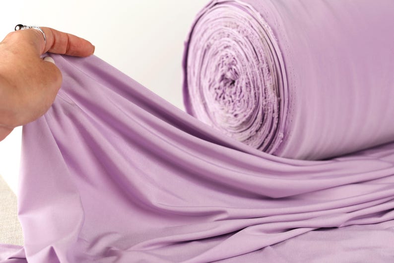 A hand is holding a piece of lavender fabric, which is rolled up and appears to be a large spool of the same fabric.