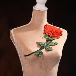 Puede incluir: Una rosa roja de tela con hojas verdes prendidas a un maniquí de tela beige.
