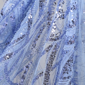 May include: Close-up of a light blue fabric with intricate sequin and bead embellishments. The fabric has a sheer, delicate appearance, with a pattern of flowing lines and scattered sequins. The sequins and beads add a shimmering effect.