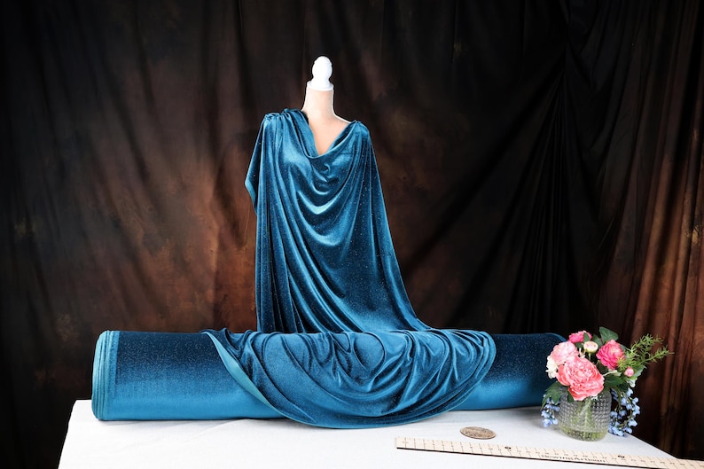 a mannequin dressed in a blue velvet gown, placed on a table with a vase of pink flowers.