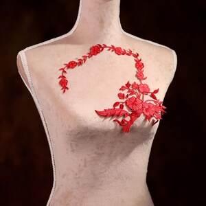 Puede incluir: Un diseño de bordado floral rojo sobre un fondo de tela blanca. El diseño presenta un grupo de flores y hojas en forma curva.