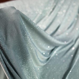 A hand is holding a piece of fabric with a shimmering, sparkly texture, which appears to be a light blue or mint-colored material.