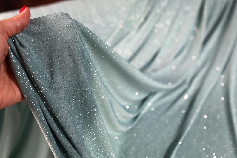 A hand is holding a piece of fabric with a shimmering, sparkly texture, which appears to be a light blue or mint-colored material.