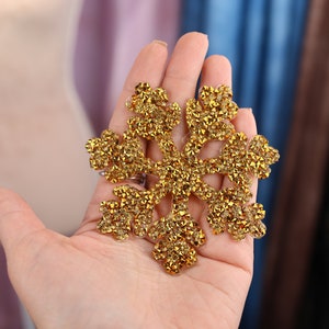 Könnte beinhalten: Ein goldener Schneeflocken-Anhänger aus kleinen, glitzernden Strasssteinen. Die Schneeflocke wird in der Hand einer Person gehalten.