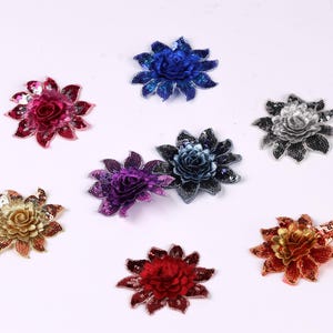 May include: A collection of decorative floral embellishments in various colors, including red, gold, blue, and purple. Each flower has a layered petal design and a central, textured core, with a sparkling, sequined appearance.