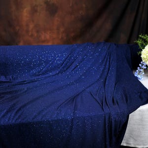 a blue fabric with a sparkly texture, draped over a white tablecloth. Next to the fabric, there is a vase filled with colorful flowers, creating a visually appealing contrast between the blue fabric and the vibrant flowers.