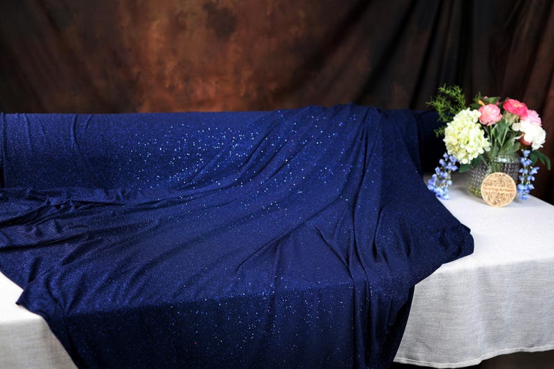 a blue fabric with a sparkly texture, draped over a white tablecloth. Next to the fabric, there is a vase filled with colorful flowers, creating a visually appealing contrast between the blue fabric and the vibrant flowers.