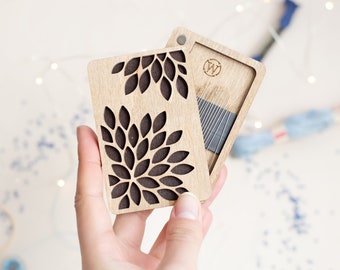 May include: A wooden needle case with a floral design. The case is open, revealing needles and a magnetic closure. The floral design is cut out of the wood, creating a decorative effect. The case is held in a hand.