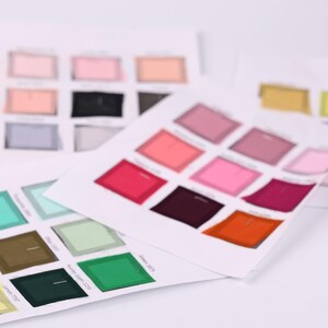 May include: Multiple white paper sheets displaying a grid of various colored fabric swatches. The swatches include shades of pink, red, orange, green, blue, yellow, and neutral tones, each labeled with small text identifying the specific color name.