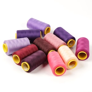 Könnte beinhalten: Eine Sammlung bunter Garnrollen in verschiedenen Lila-, Rosa- und Burgundertönen. Die Spulen haben gelbe Kerne und sind auf einer weißen Oberfläche angeordnet. Ideal für Näh- und Bastelprojekte.