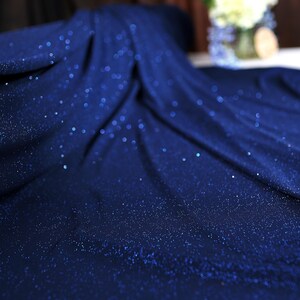 a close-up view of a blue fabric with a sparkling texture, possibly made of sequins or glitter. The fabric appears to be folded or draped, creating a sense of movement and depth. In the background, there is a vase with flowers, adding a touch of natural beauty to the scene.