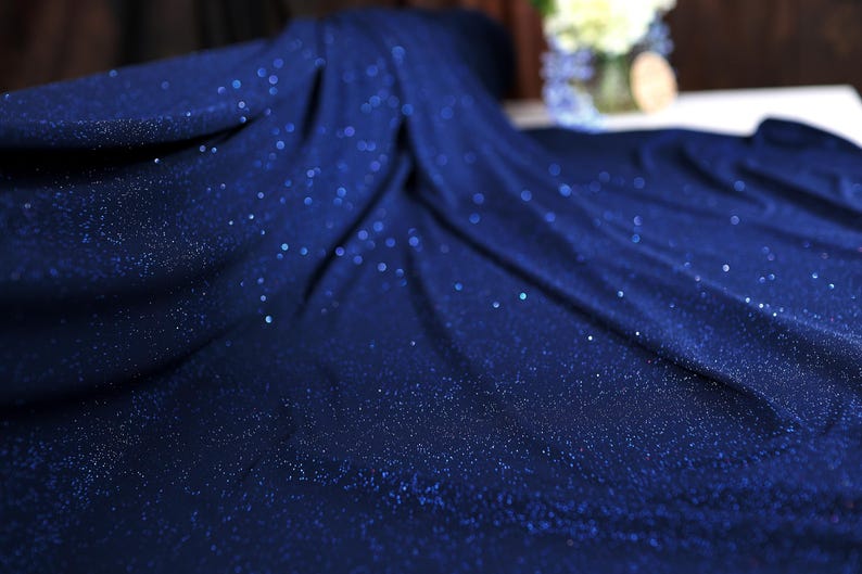 a close-up view of a blue fabric with a sparkling texture, possibly made of sequins or glitter. The fabric appears to be folded or draped, creating a sense of movement and depth. In the background, there is a vase with flowers, adding a touch of natural beauty to the scene.