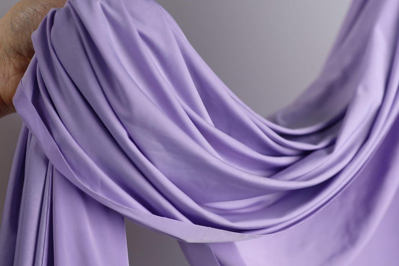 A hand is holding a piece of lavender fabric, which appears to be a soft, flowing material.