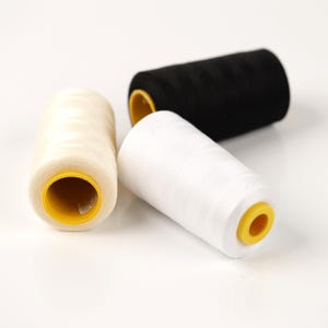 May include: Three spools of thread in cream, white, and black. The spools have yellow centers. The threads are tightly wound, and the image is well-lit, highlighting the texture of the thread.