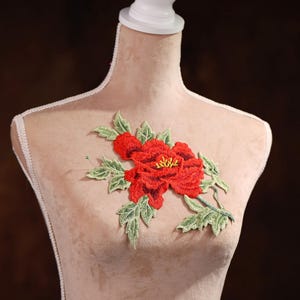 Puede incluir: Una flor roja bordada con hojas verdes sobre un fondo de tela beige.