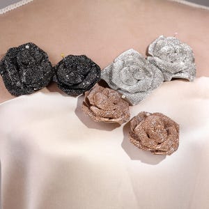 May include: An assortment of sparkling, beaded rose brooches in various colors, including black, silver, and rose gold. The brooches are arranged on a cream-colored fabric, showcasing their intricate details and shimmering surfaces. These decorative accessories add a touch of elegance.